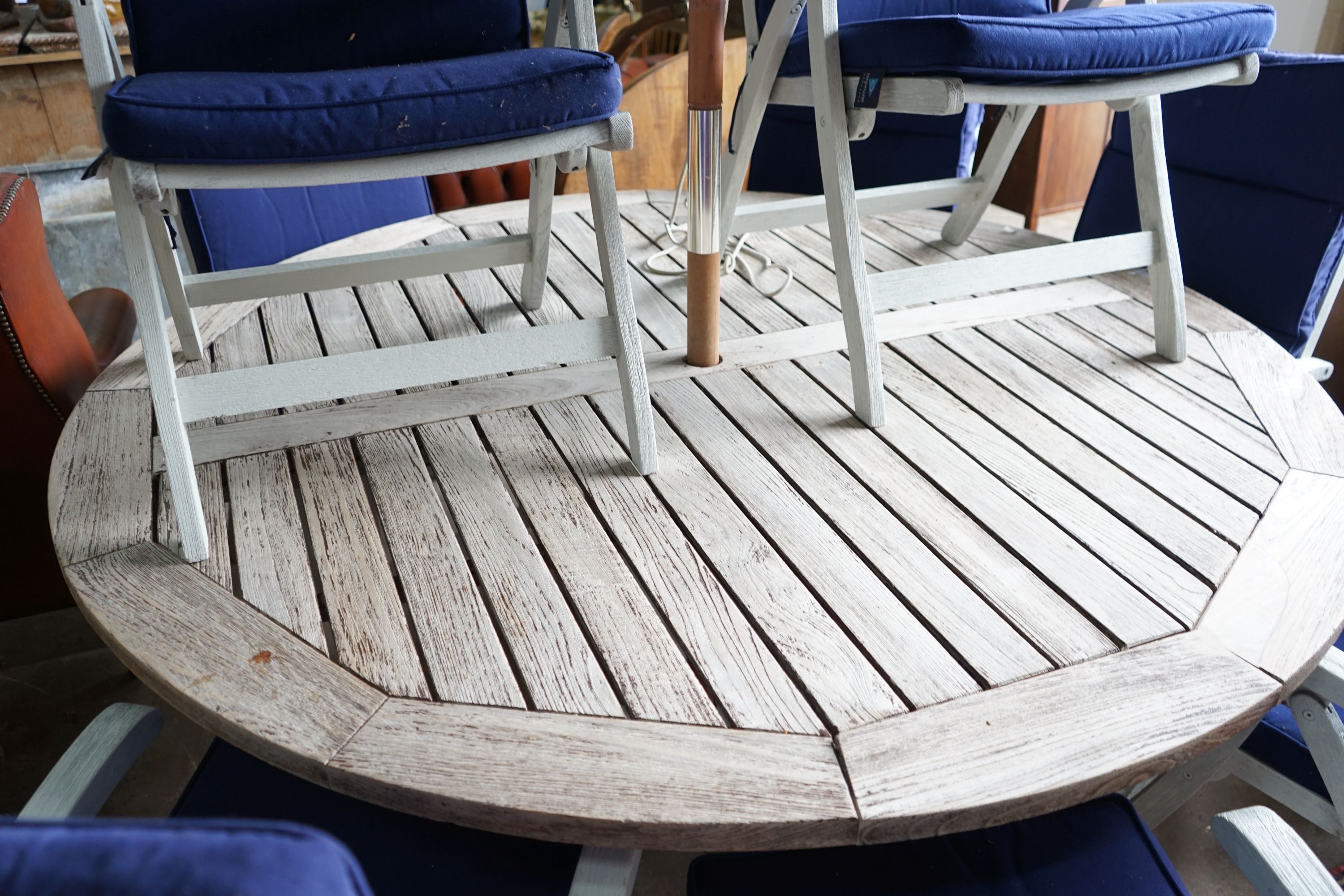 A large circular weathered teak garden table, diameter 180cm, height 72cm, eight painted teak folding chairs with seat pads and a blue canvas crank parasol